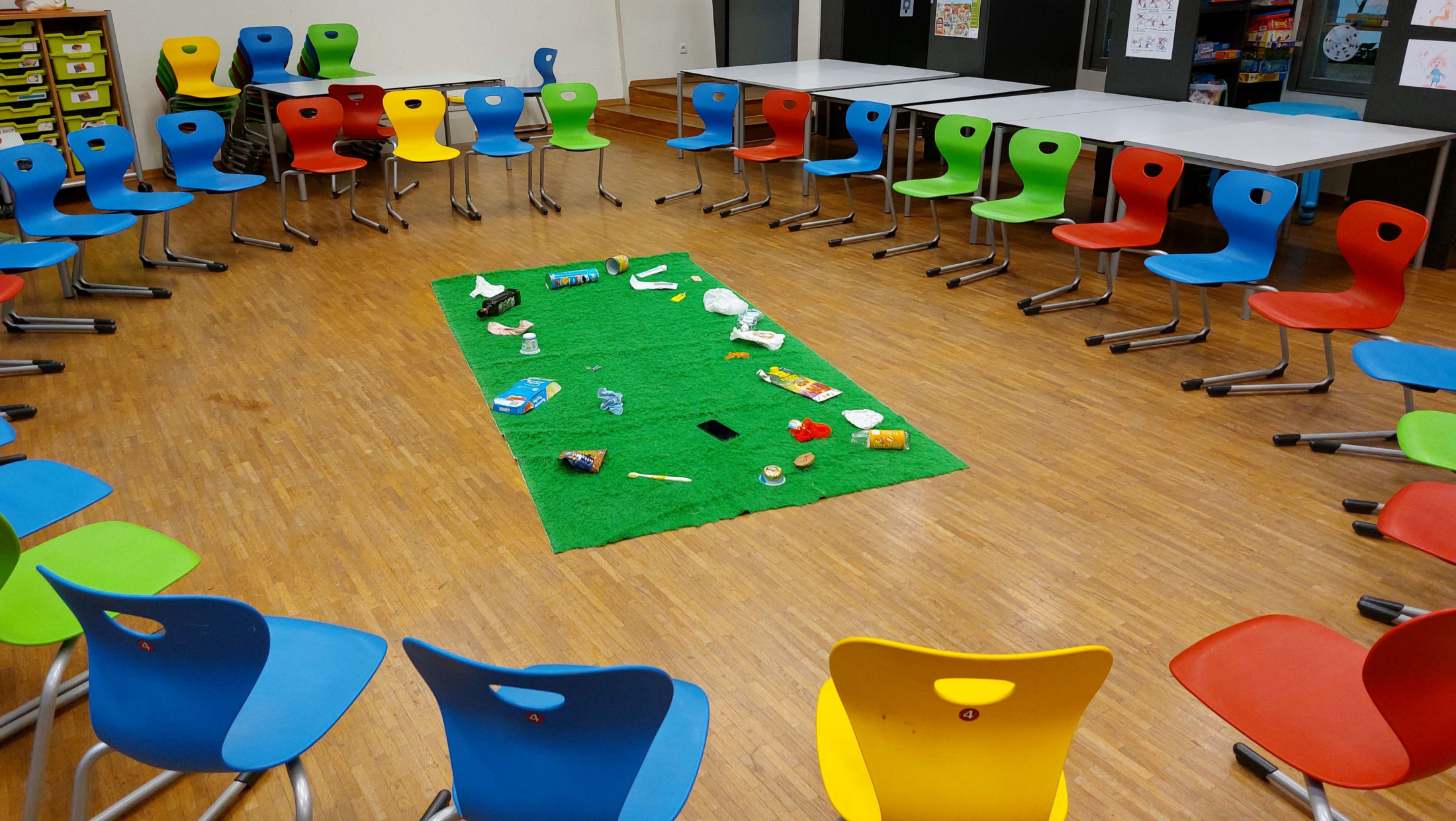 Ein Klassenzimmer mit Stühlen im Kreis um eine grüne Matte mit Müll. Tische im Hintergrund.