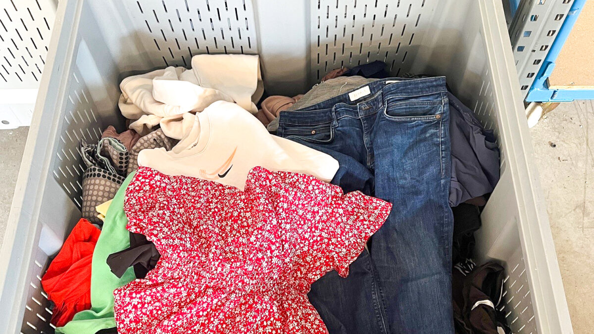 Ein grauer Behälter voller bunter Kleidung, darunter Jeans und ein rotes Blumenkleid.