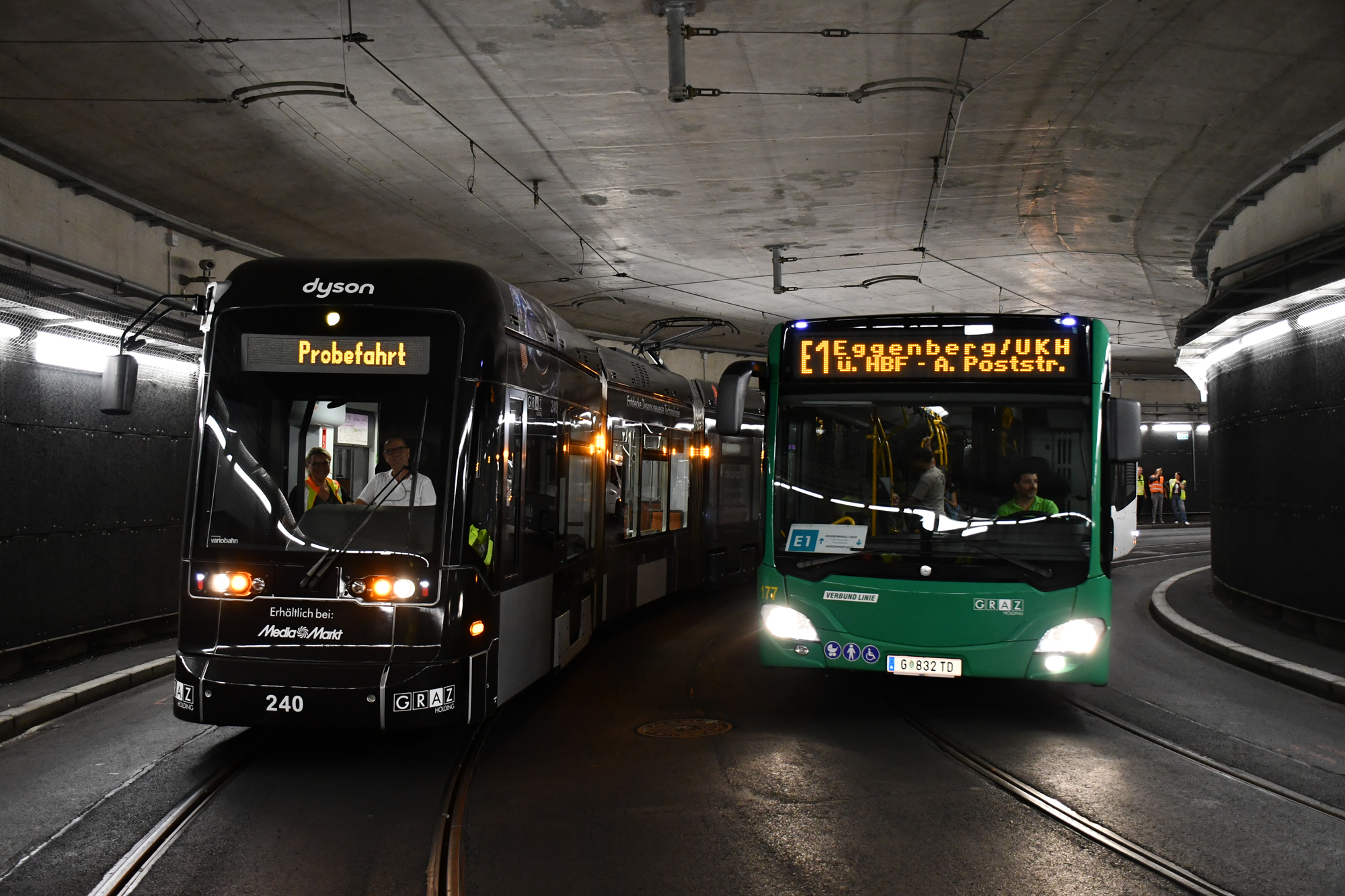 Schwarze Dyson Straßenbahn "Probefahrt" und grüner Bus E1 Graz in Tunnel.