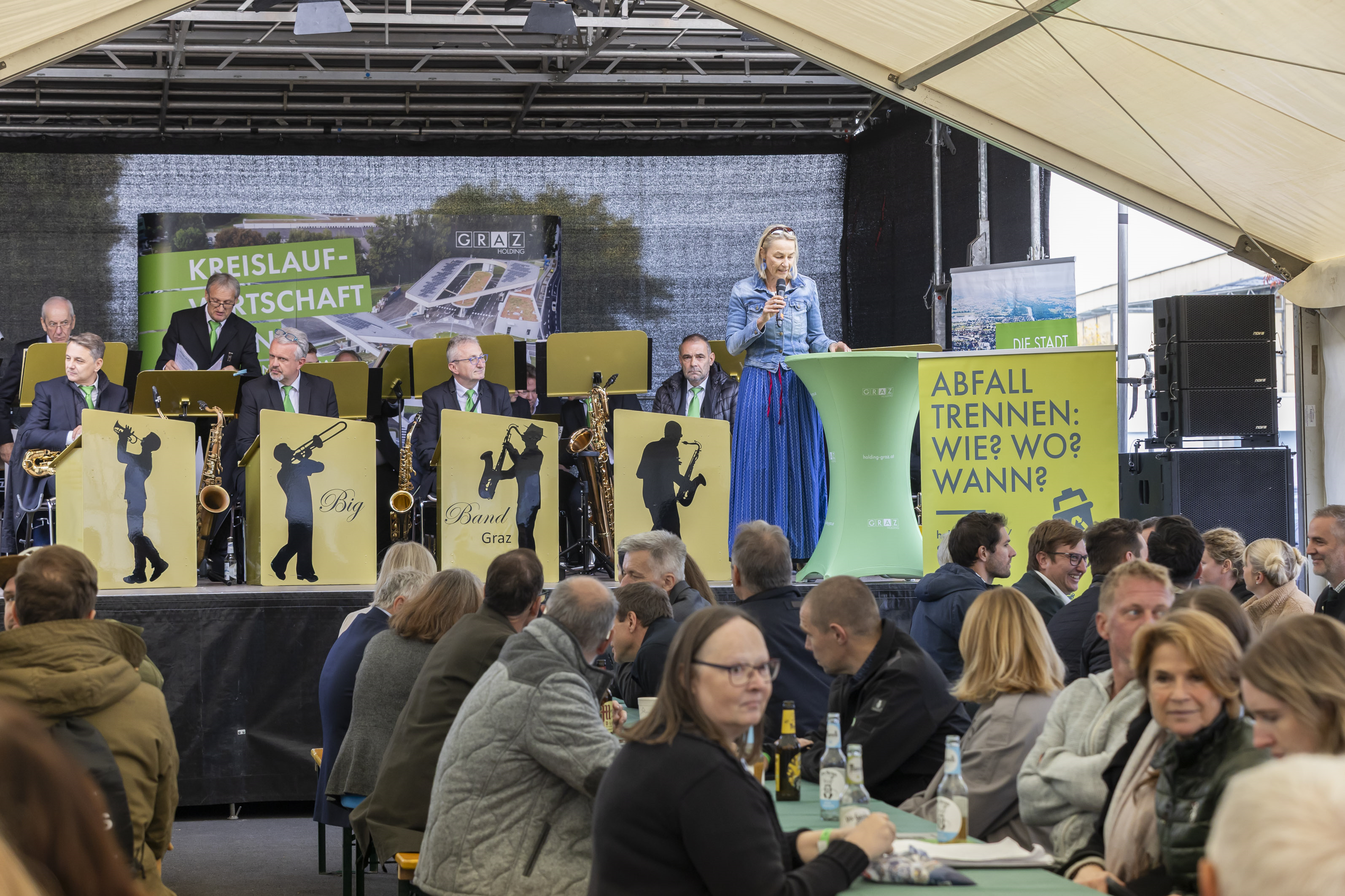 Eine Frau spricht auf der Bühne in Graz, mit Band und Publikum.