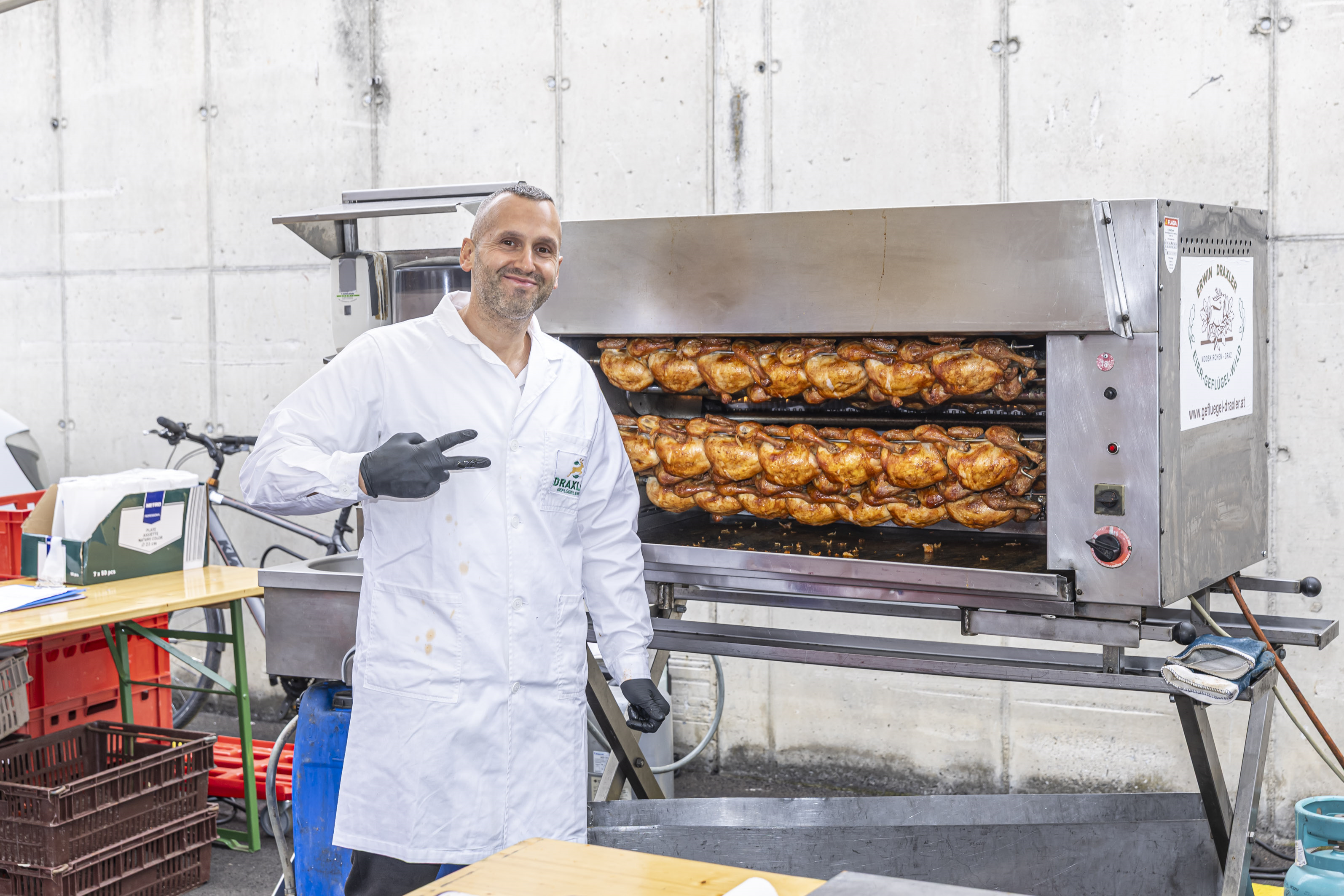 Mann mit Drehgrill-Hühnern in Graz, weiβer Kittel, Friedenszeichen.
