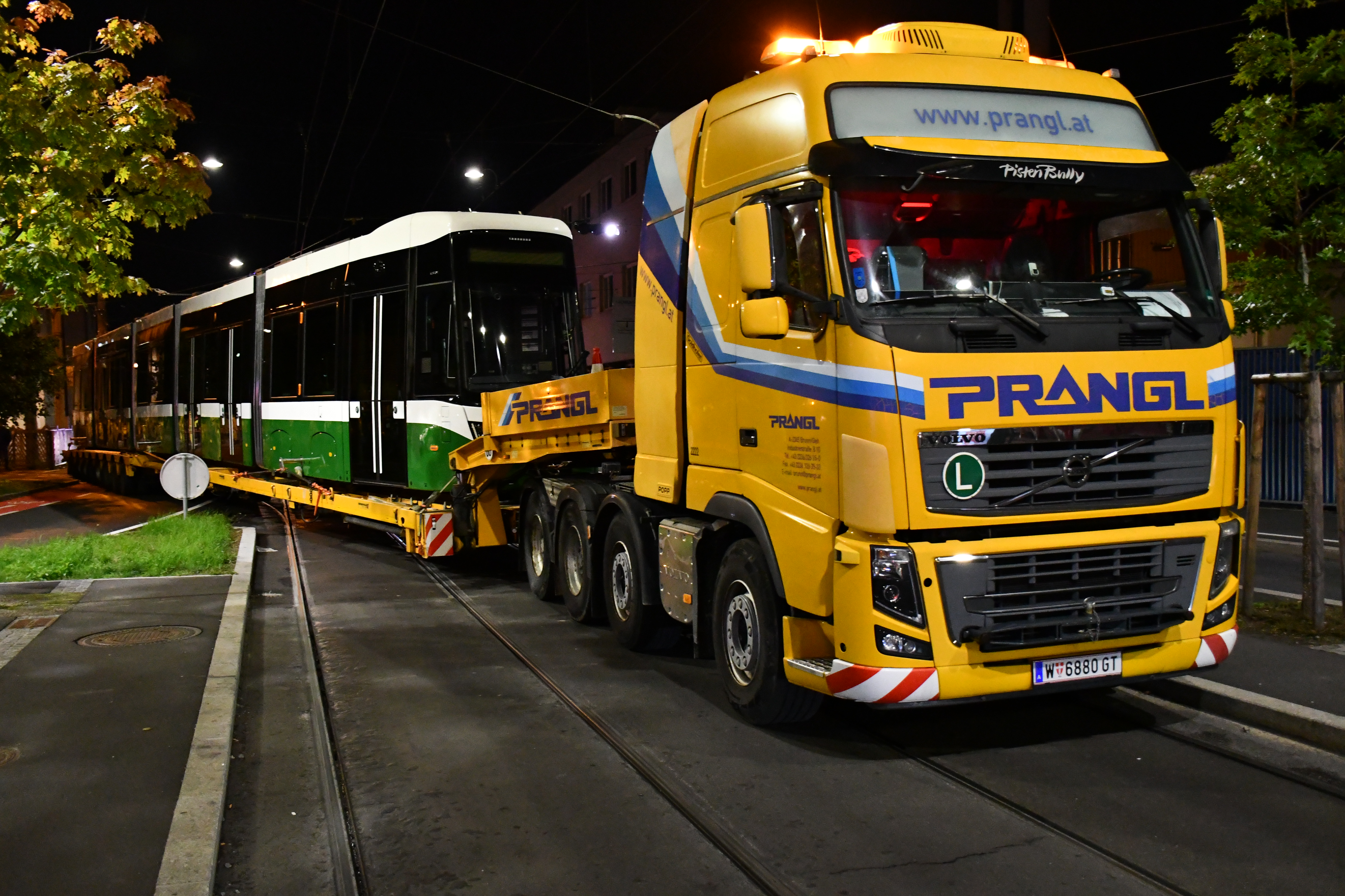 Gelber Prangl-LKW transportiert eine grüne Straßenbahn in Graz bei Nacht.