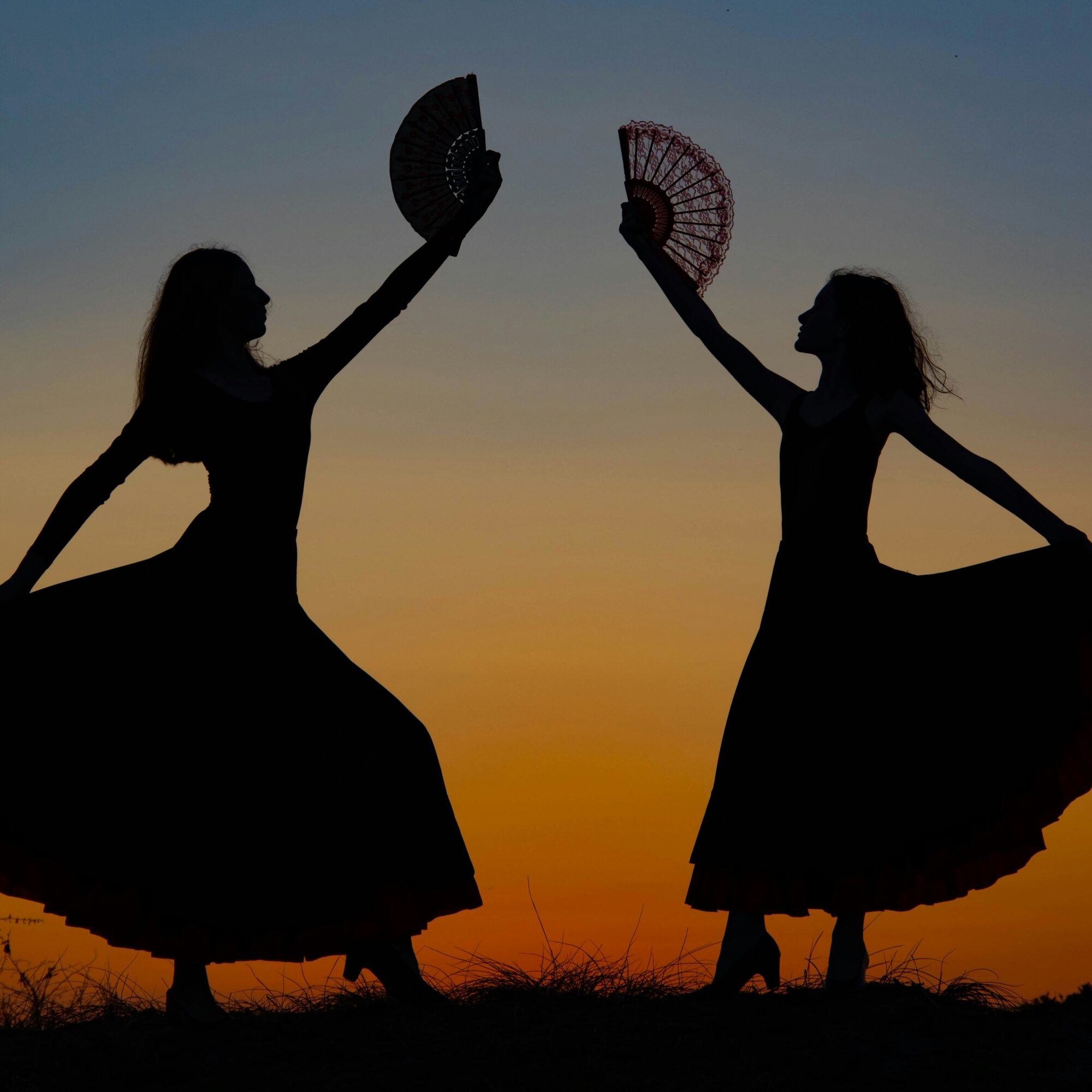 Zwei tanzende Frauensilhouetten mit Fächern vor Sonnenuntergang.