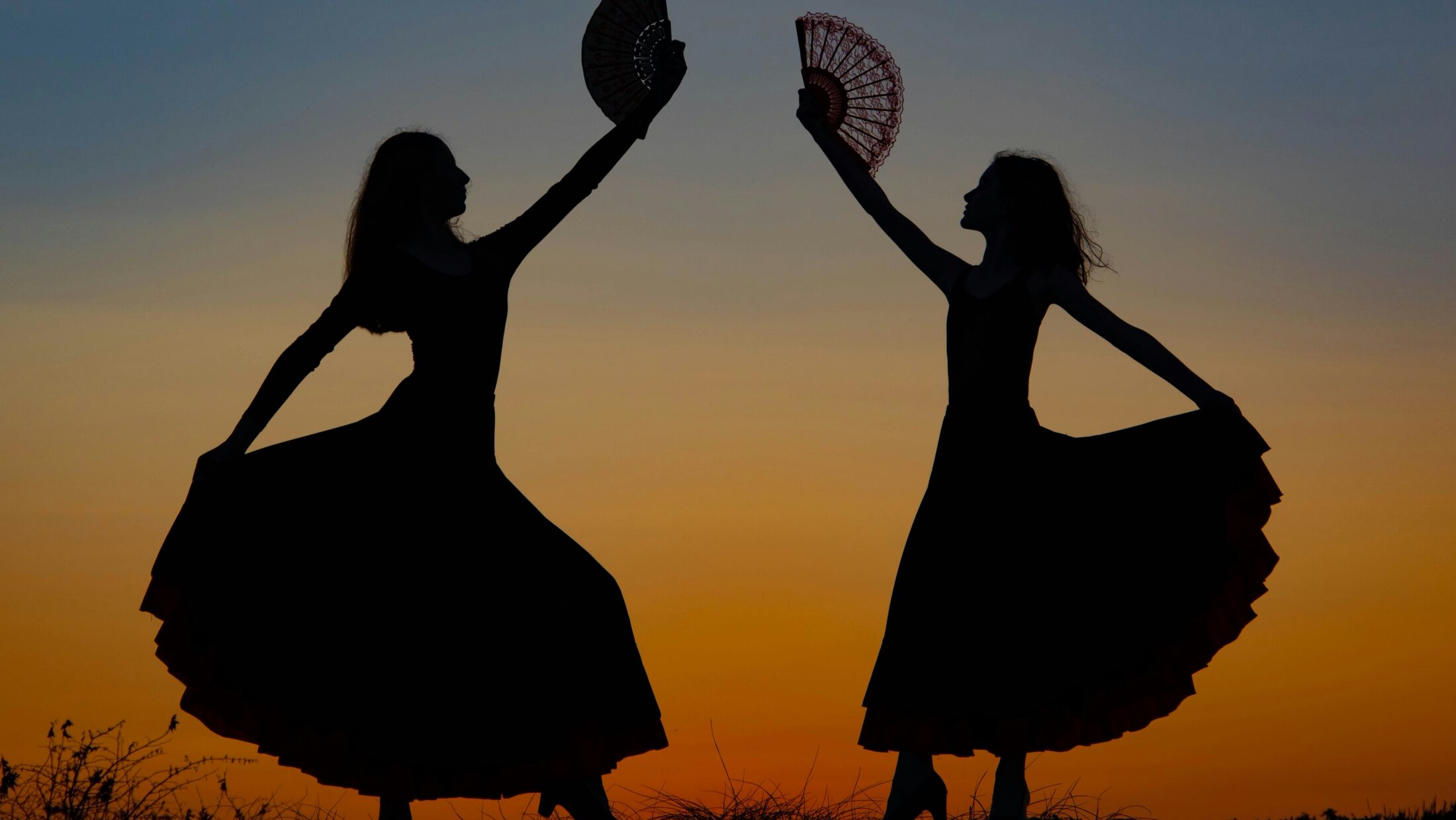 Zwei tanzende Frauensilhouetten mit Fächern vor Sonnenuntergang.