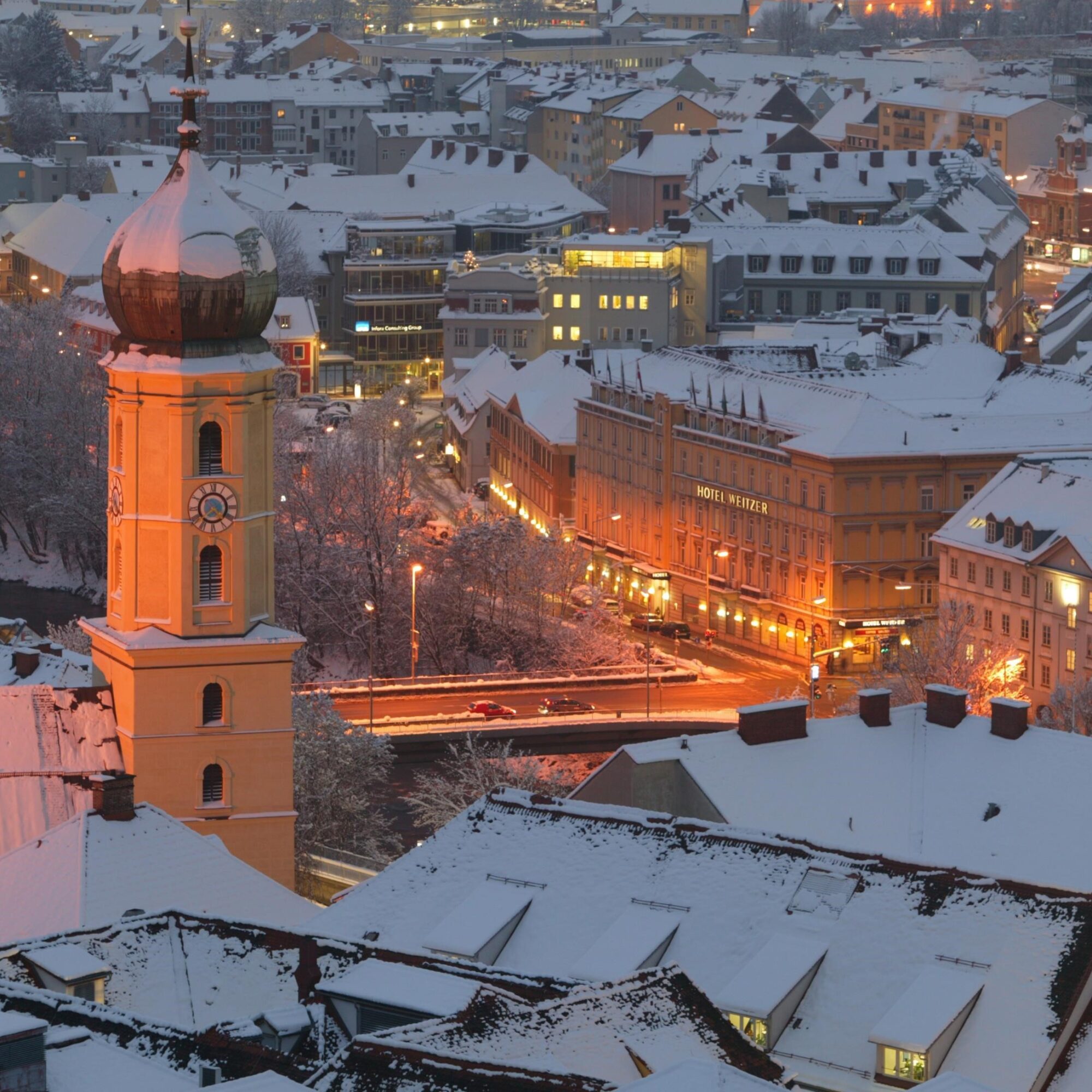 Winterliche Luftaufnahme von Graz mit schneebedeckten Dächern und Kirchturm.