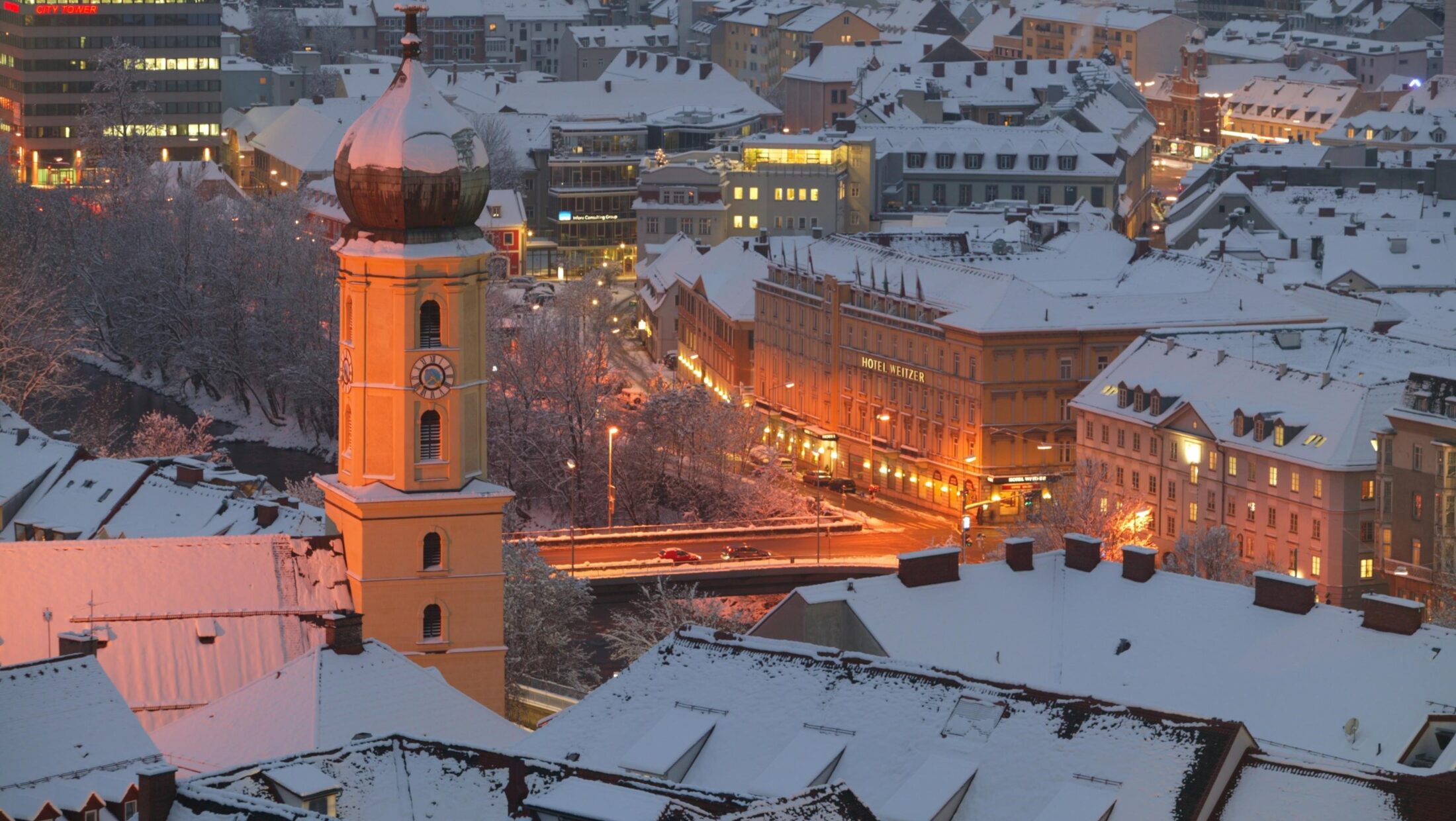 Winterliche Luftaufnahme von Graz mit schneebedeckten Dächern und Kirchturm.