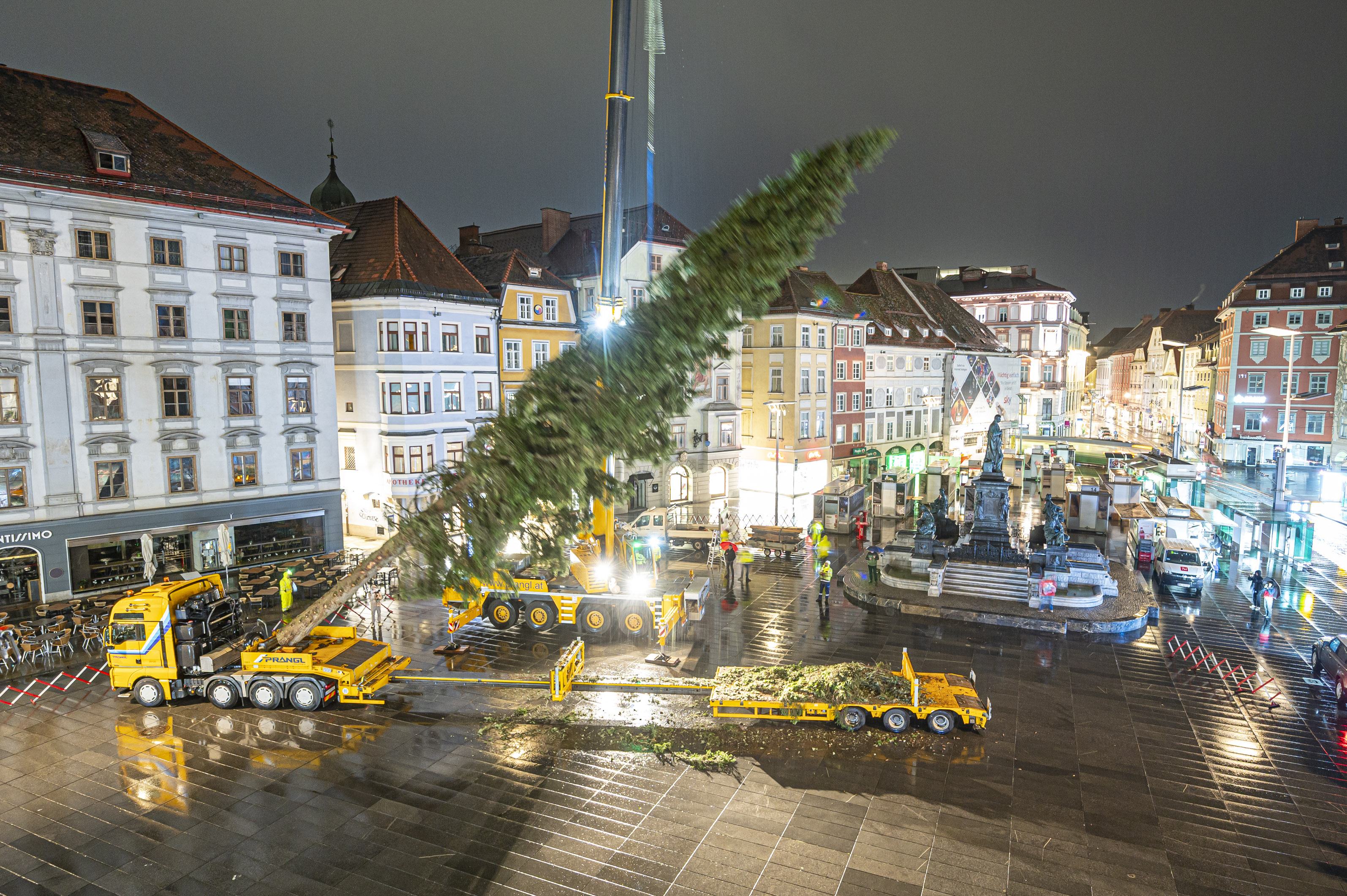 Ein Kran hebt einen Weihnachtsbaum in Graz auf einem regnerischen Platz.