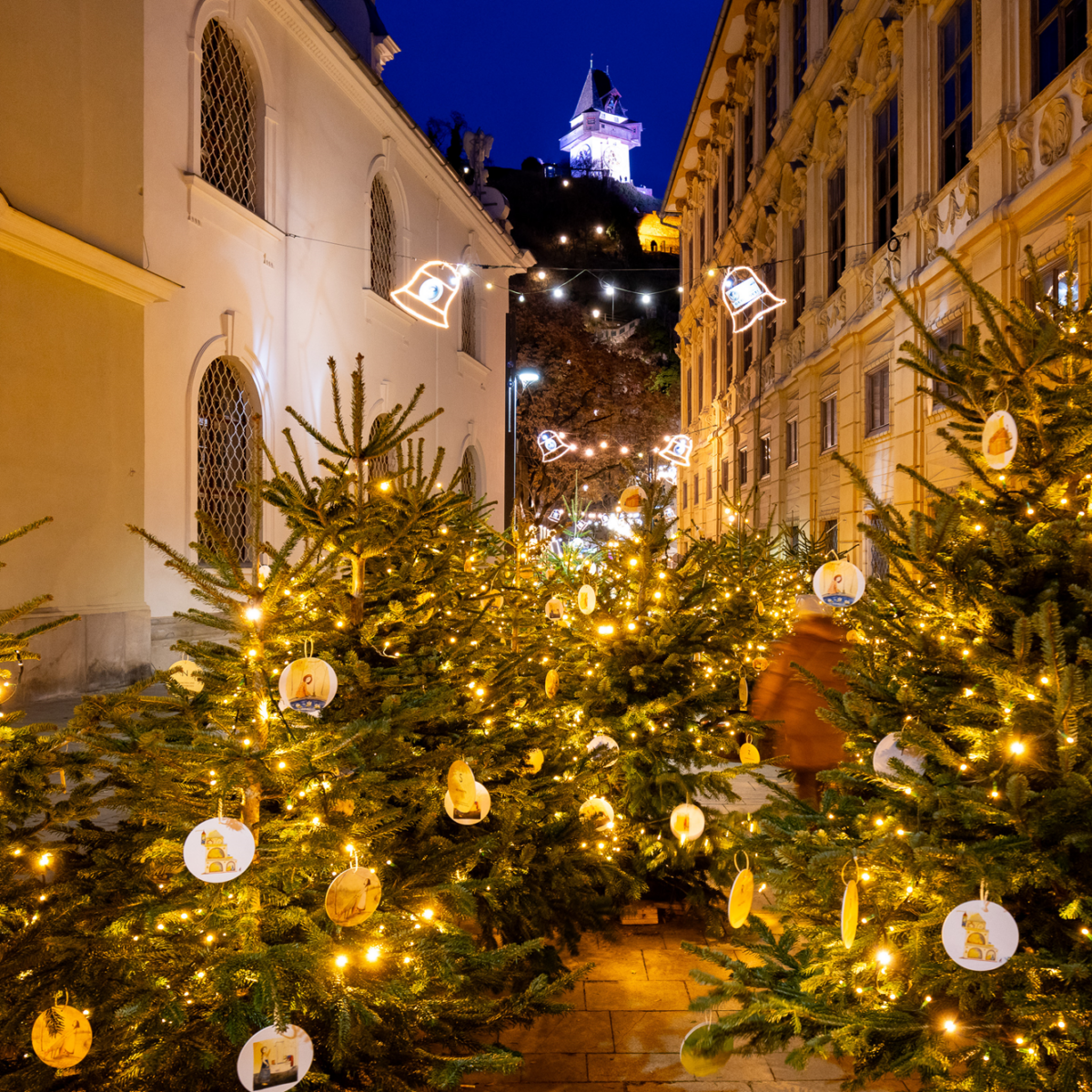 Weihnachtsmarkt Graz mit Christbäumen, Lichtern und Glocken. Grazer Uhrturm im Hintergrund.