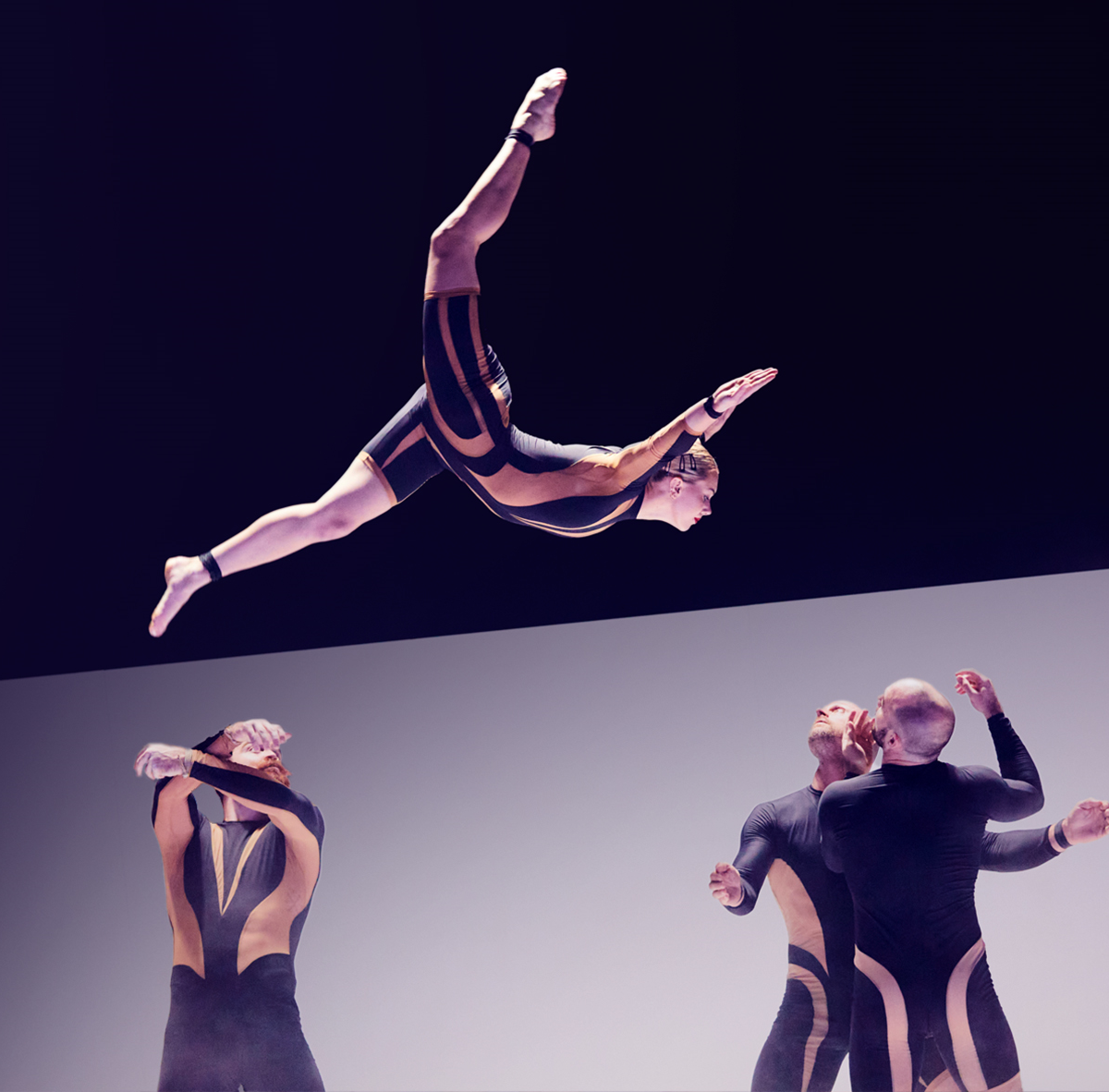 Eine Frau springt, gehoben von drei Männern in engen Anzügen.