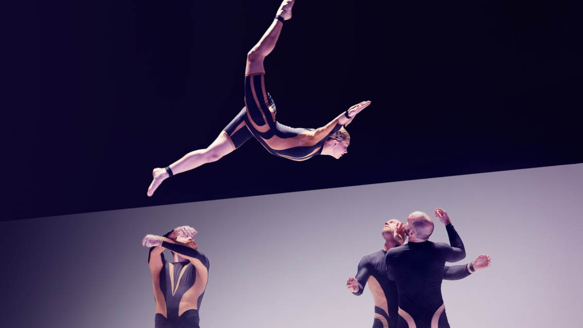 Eine Frau springt, gehoben von drei Männern in engen Anzügen.