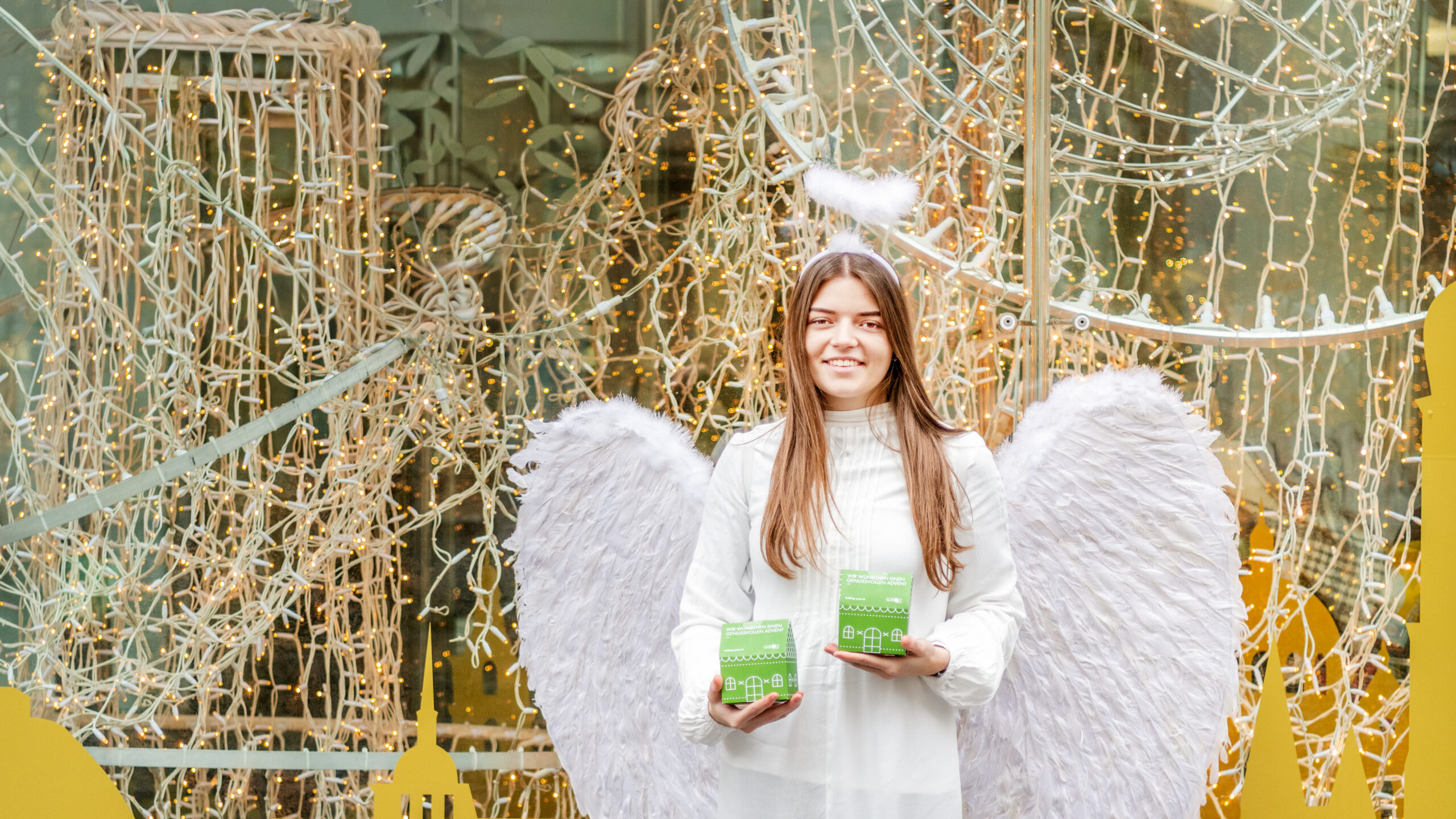 Ein Engel mit Heiligenschein und Flügeln hält grüne Schachteln vor einem beleuchteten Schaufenster in Graz.