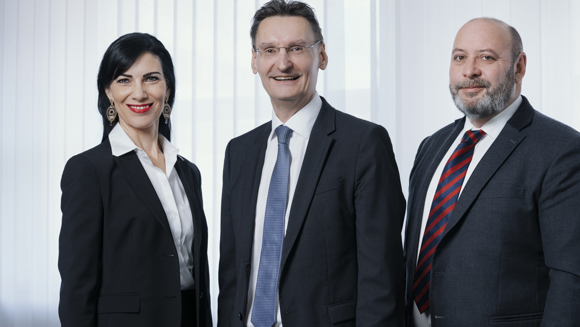 Drei Personen in Anzügen vor hellem Hintergrund.