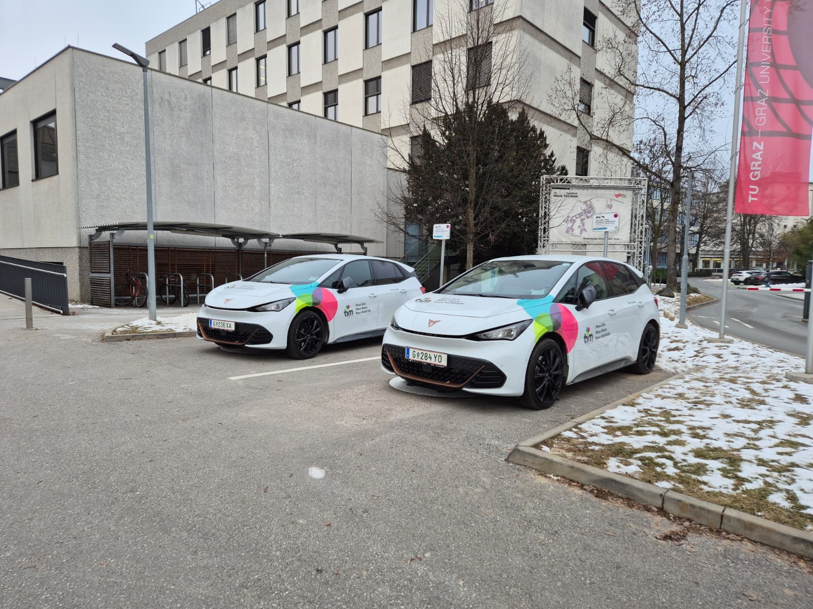 Zwei weiße Autos vor der TU Graz geparkt, mit Logos versehen.